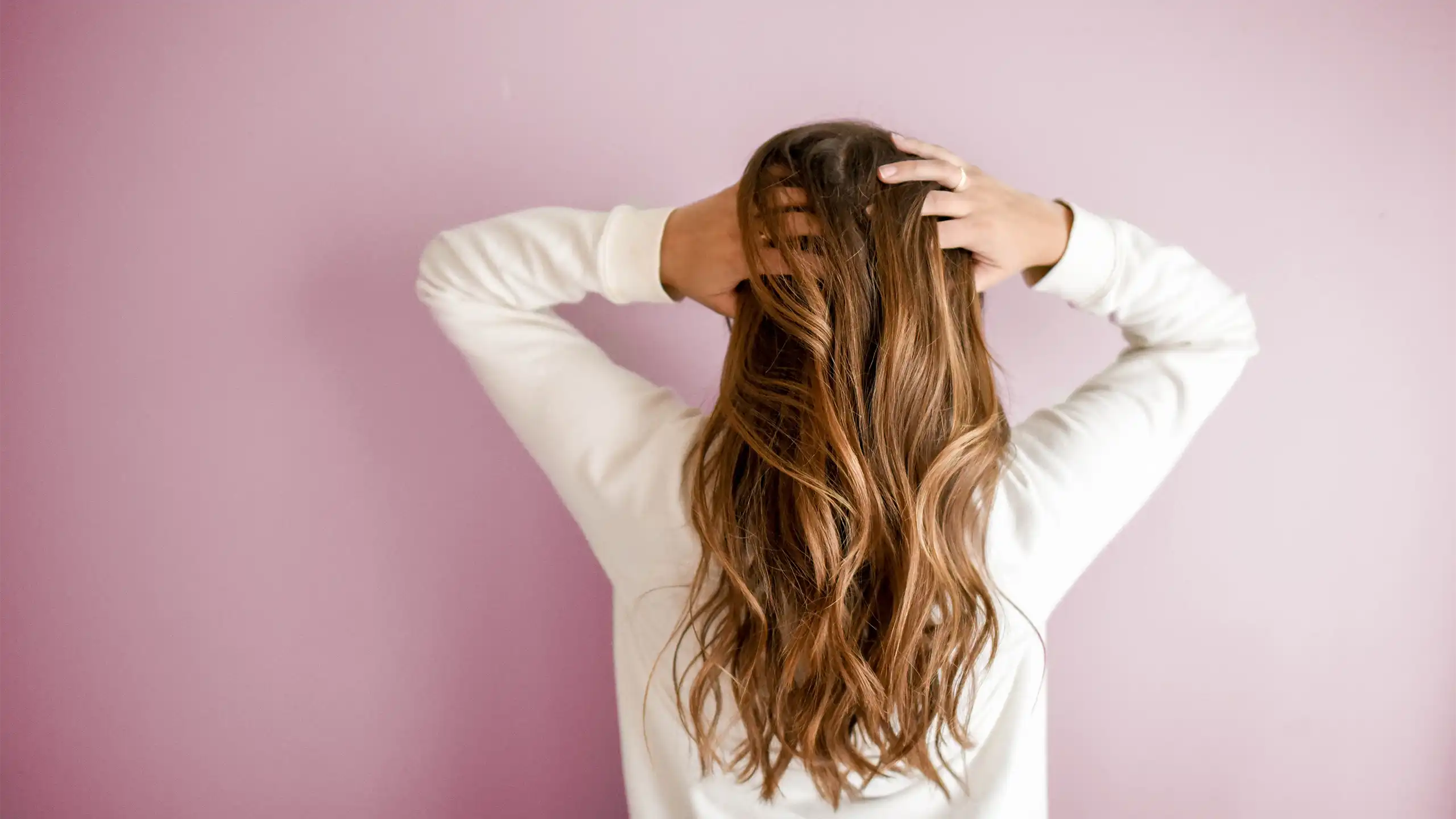 A woman holding the back of her head, long hair flowing down her back, against a pink wall.